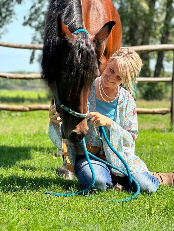 Tammy kneeling in the grass with her horse lowering its head to meet her