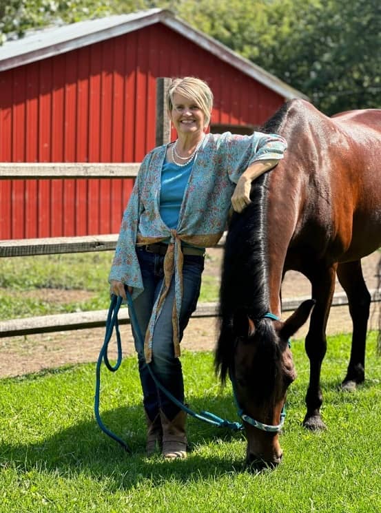 Rider standing beside her grazing horse by a red barn
